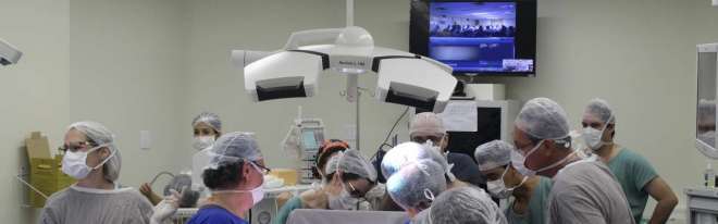 Duas equipes médicas com 20 profissionais participaram da cirurgia. TV ao fundo mostra os 50 médicos e profissionais de saúde que acompanharam o procedimento via teleconferência