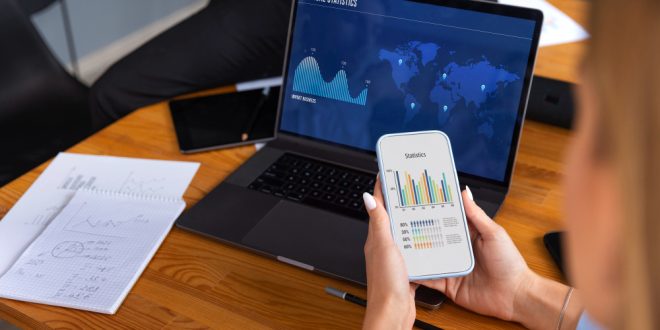 Uma pessoa usando notebook e celular enquanto analisa gráficos financeiros